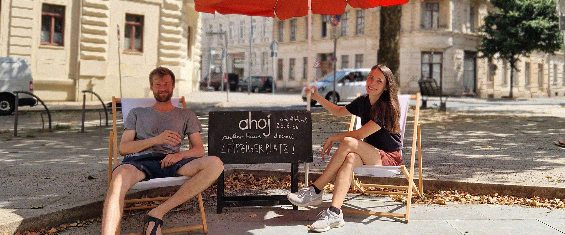 Zwei Personen sitzen auf Liegestühlen am Leipziger Platz in Görlitz neben einer AHOJ-Tafel.