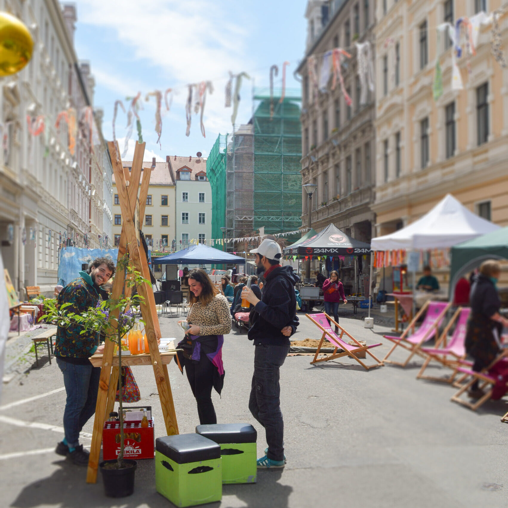 Straßenfest in der Görlitzer Innenstadt mit Marktständen, bunten Stoffbändern, Besucher*innen an einem Stand und Menschen in Liegestühlen.